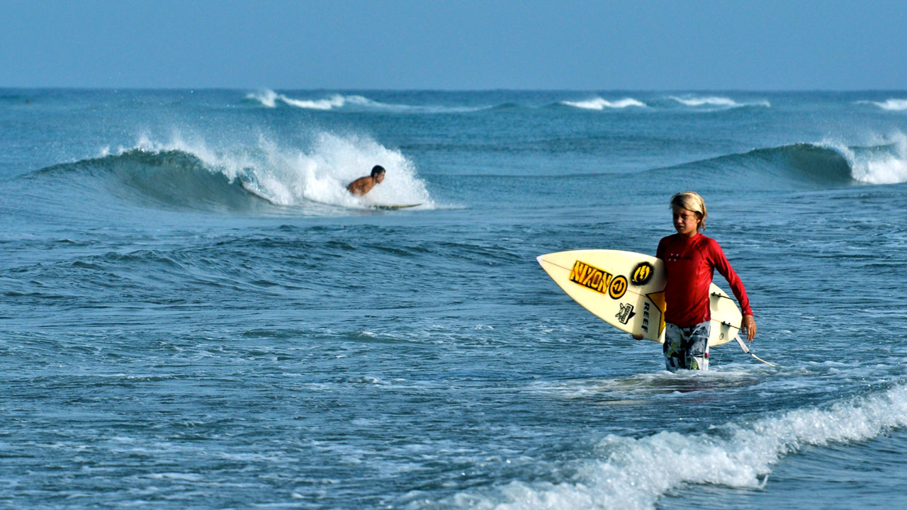 Niño con tabla de surf bajo el brazo, camina por la orilla